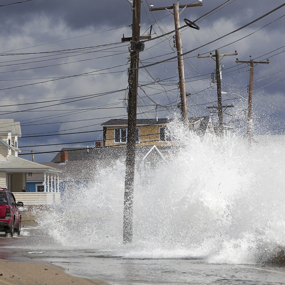Waves crash against a break wall in conjunction with high tide and a storm as a red car drives on the road.