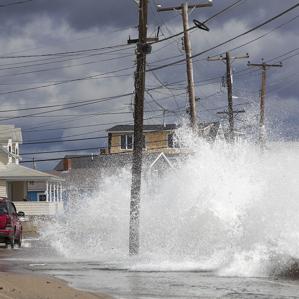 coche rojo inundando el océano Waves crash against a break wall in conjunction with high tide and a storm as a red car drives on the road.